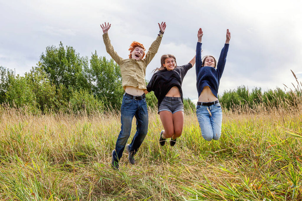 Summer holidays vacation happy people concept. Group of three friends boy and two girls jumping, dancing and having fun together outdoors. Picnic with friends on road trip in nature - Photo, Image
