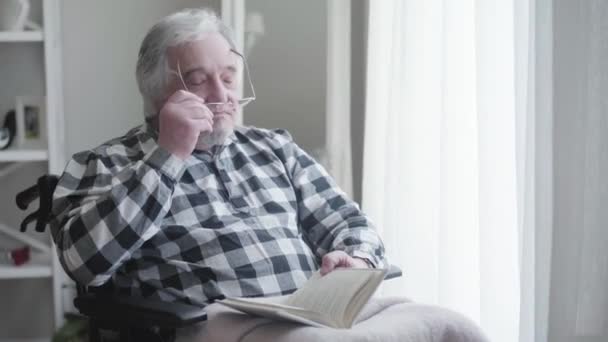 Portrait of elderly Caucasian man putting on eyeglasses and reading book. Disabled old retiree sitting in wheelchair at home on sunny day. Disability, retirement, medical problems. - Filmagem, Vídeo