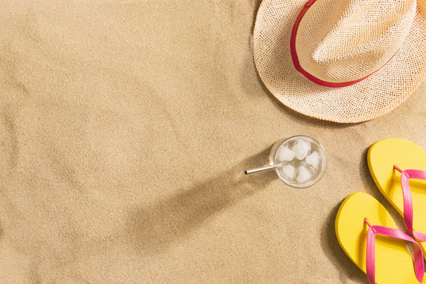 Summer vacation composition. Hat, glass of water with ice and flip flops on sand background. Harsh light with shadows. Summer background. - Photo, image