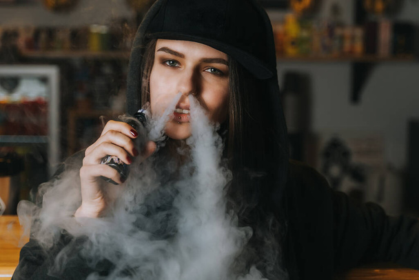 Young pretty woman in cap smoke an electronic cigarette at the vape shop. Hip-hop style. Closeup. - Foto, imagen