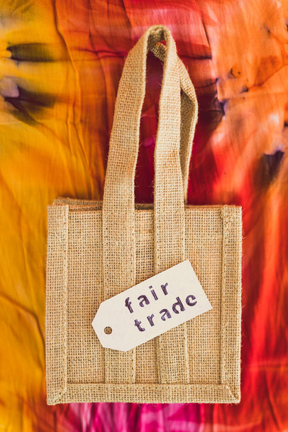 ethical shopping and locally sourced items conceptual still-life, small canvas bag made of natural materials with Fair Trade text on price tag on hippie tie dye background - Zdjęcie, obraz