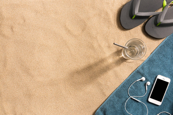 Summer vacation composition. Flip flops, smartphone and glass of water on sand background. Harsh light with shadows. Summer background. Border composition made of towel - Фото, изображение