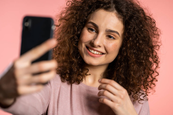 Smiling happy girl with curly hair making selfie on smartphone over pink background. Technology, mobile device, social networks concept. - Фото, зображення