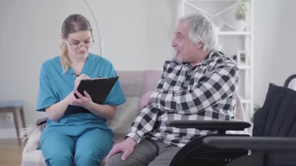 Professional Caucasian nurse talking with elderly male patient and writing down symptoms. Elderly man and adult woman sitting on couch in nursing home. Communication, treatment, disability, help. - 映像、動画