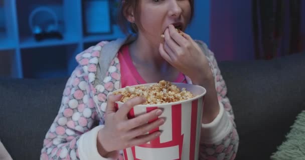 Young woman sitting on the sofa in the living room eating popcorn watching TV - Filmagem, Vídeo
