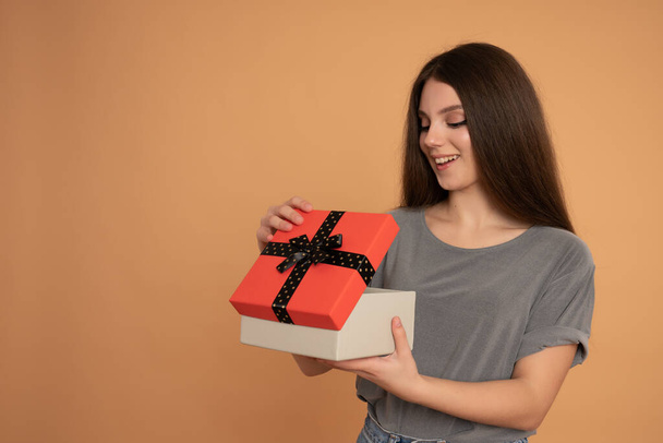 Glad young woman opening her birthday gift. Studio portrait of beautiful brunette teen girl with new year present. - 写真・画像