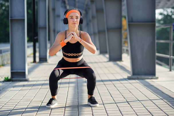 Fitness girl with earphones which doing squat exercises with fabric booty band during her sport workout on special sportground - Φωτογραφία, εικόνα