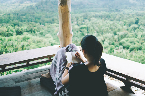 Frau schreibt in kleines weißes Notizbuch, um sich Notizen zu machen, nicht zu vergessen, zu planen oder ein Buch zu schreiben mit schöner Terrasse des Hauses und Natur-Bergblick Hintergrund. - Foto, Bild