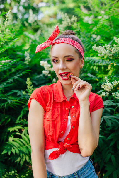 Bela jovem pin up menina vestida de camisa vermelha com maquiagem brilhante comendo morango natural fresco
. - Foto, Imagem