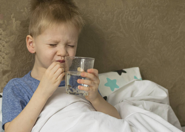 Een ziek, verdrietig kind met koorts en hoofdpijn ligt naast medicijnen en pillen in bed. Griep verkoudheid virus bacterie - Foto, afbeelding