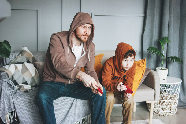 Father and son tween boy in hoodies playing computer games with joystick at the home tipical caucasian family - Photo, Image