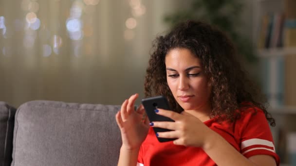 Serious arab girl in red checking smart phone sitting on a couch in the living room at home  - Imágenes, Vídeo
