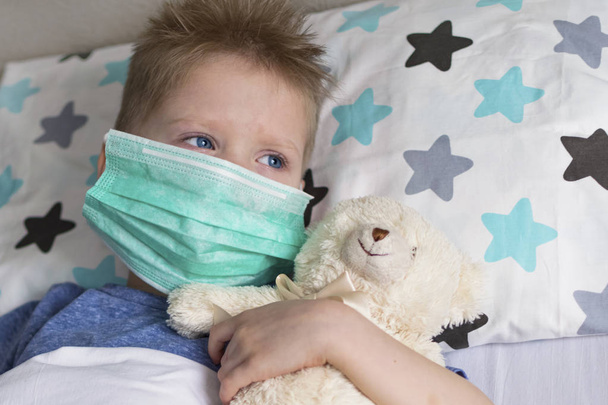 Un enfant malade et triste dans un masque avec une température et un mal de tête se trouve au lit. Grippe rhume maladie virus bactérie
 - Photo, image