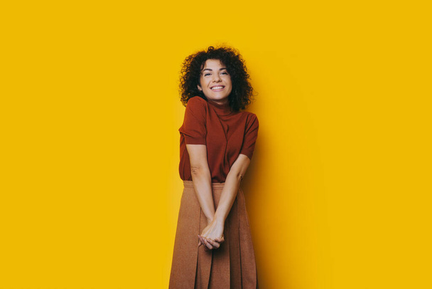 Preciosa chica caucásica con el pelo rizado y sonrisa alegre posando en un bonito vestido con las manos cruzadas sobre un fondo amarillo
 - Foto, Imagen