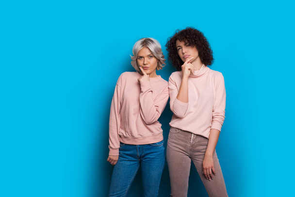 Two caucasian sisters dressed in pink sweaters and jeans are looking thoughtful posing on a blue background - Photo, Image