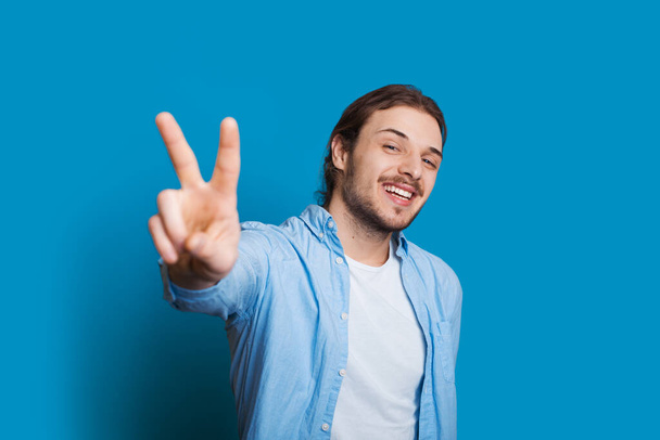 Усміхнений незахищений білий чоловік з довгим волоссям, одягнений у футболку і блакитна сорочка жестикулює знак перемоги синій фон
 - Фото, зображення