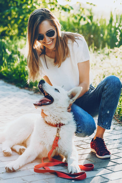 Lächelndes kaukasisches Mädchen in blauen Jeans und weißem T-Shirt spielt fröhlich mit ihrem Hund im Licht der Sonne - Foto, Bild