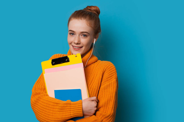 Cute smiling student with red hair and freckles is holding some books being dressed in an orange sweater while posing on a blue background and listening to - Photo, Image