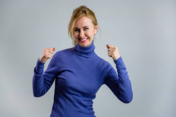 Happy smiling blonde woman in a blue sweater on a blue background shows the joy of good luck - Zdjęcie, obraz