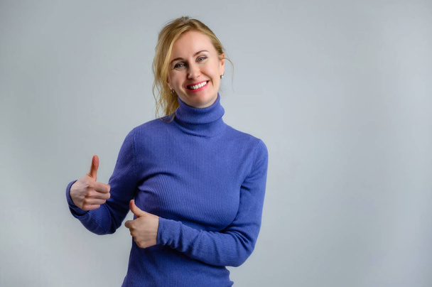 Happy blonde woman with a smile in a blue sweater on a blue background shows ok - Foto, immagini