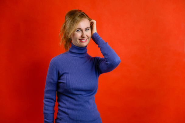 Portrait of a smiling blonde woman in a blue sweater on a red background shows happiness and joy - Фото, зображення