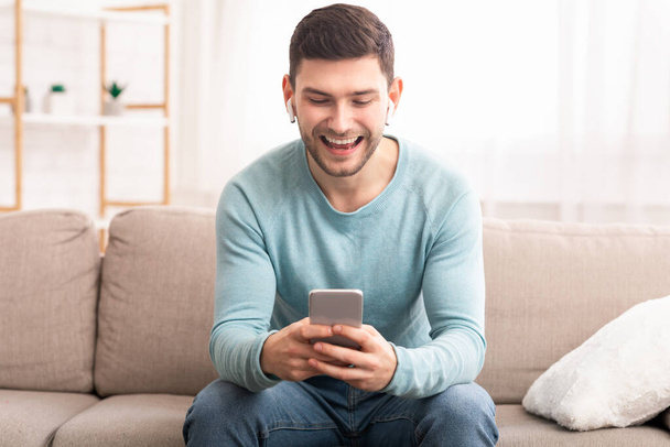 Guy In Earbuds Using Phone Sitting On Couch At Home - Foto, imagen