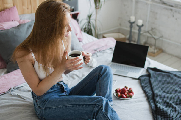 Blonde sexy woman with a cup of tea or coffee and strawberries having fun in bed. She wearing white bra and casual jeans. Happy morning - Foto, Imagem