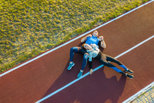 夏のジョギングマラソンの後に休憩スタジアムフィールドの赤いゴム走路に横たわっている2人の若いスポーツマンとスポーツ女性の空中ビューをトップダウン. - 写真・画像