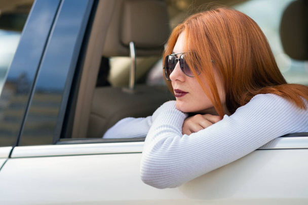 Jeune femme aux cheveux roux et lunettes de soleil voyageant en voiture. Passager regardant par la fenêtre arrière d'un taxi dans une ville
. - Photo, image
