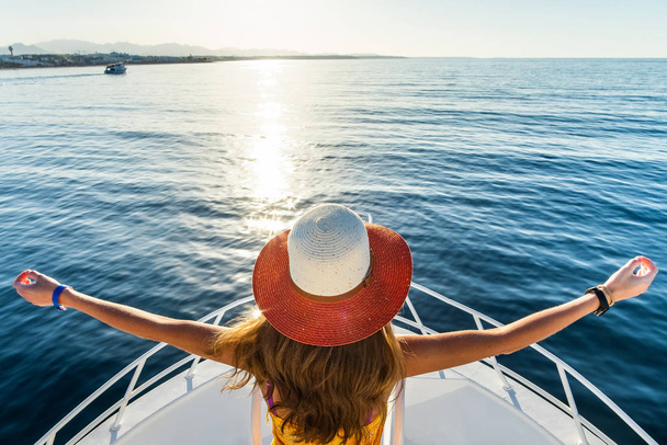 Junge Frau mit langen Haaren in gelbem Kleid und Strohhut steht mit erhobenen Händen auf weißem Jachtdeck und genießt Blick auf blaues Meerwasser. - Foto, Bild