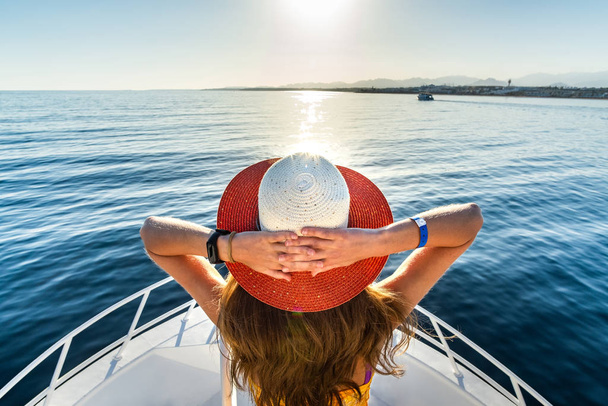 Jeune femme aux cheveux longs portant une robe jaune et un chapeau de paille debout sur le pont de yacht blanc bénéficiant d'une vue sur l'eau de mer bleue. - Photo, image