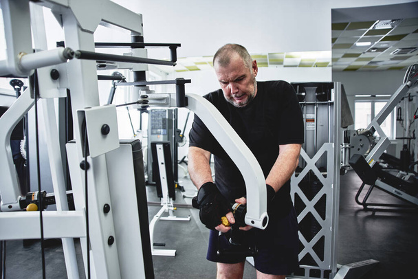 Adult gray-haired man trains on fitness equipment in the gym, pumps legs and arms muscles, loses weight. Concept of healthy lifestyle in old age - Photo, Image