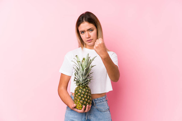 Jeune femme caucasienne tenant un ananas montrant poing à caméra, expression faciale agressive
. - Photo, image