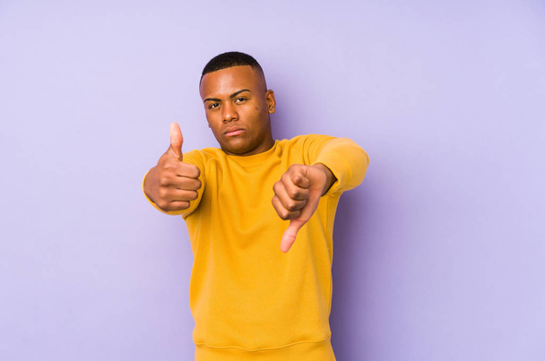 Jonge latijn man geïsoleerd op paarse achtergrond tonen duimen omhoog en duimen omlaag, moeilijk kiezen concept - Foto, afbeelding