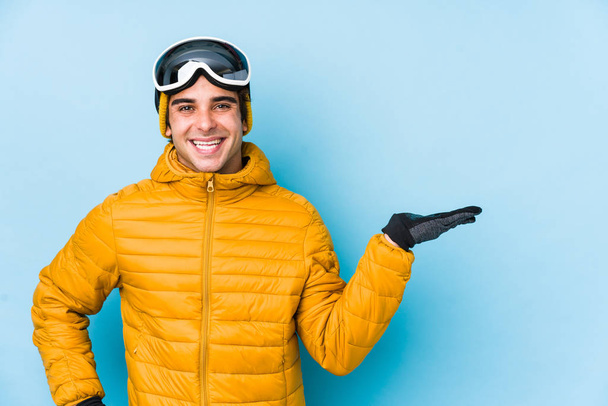 Молодой лыжник в изолированных сноубордистских очках показывает пространство для копирования на ладони и держит другую руку на талии.
. - Фото, изображение