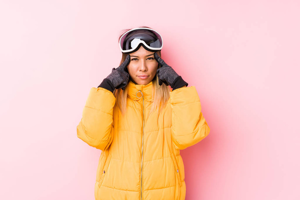 若い白人女性はタスクに焦点を当てたピンクの背景にスキー服を着て、額を頭を指して保つ. - 写真・画像