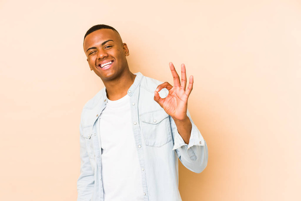 Junger lateinischer Mann auf beigem Hintergrund zwinkert und hält eine Okay-Geste mit der Hand. - Foto, Bild