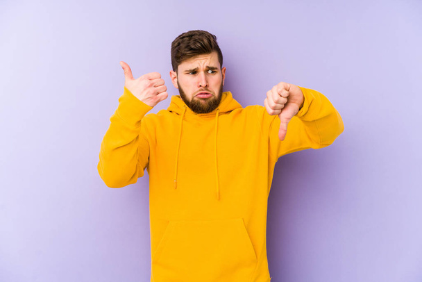 Mladý muž izolovaný na fialovém pozadí ukazující palce nahoru a palce dolů, obtížně zvolitelný koncept - Fotografie, Obrázek