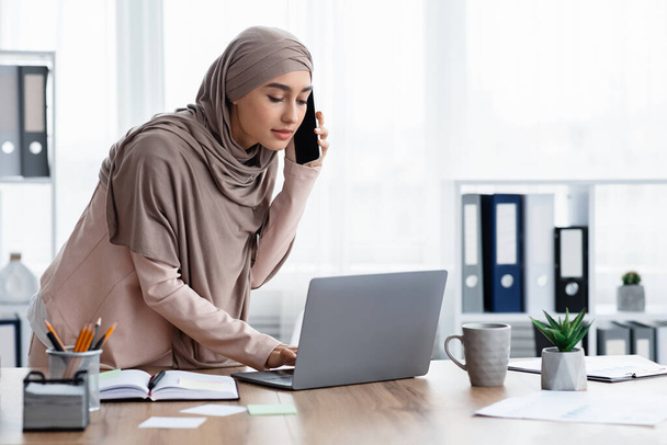 Ocupada mujer de negocios árabe utilizando el ordenador portátil y hablando por teléfono celular, consultando al cliente
 - Foto, imagen