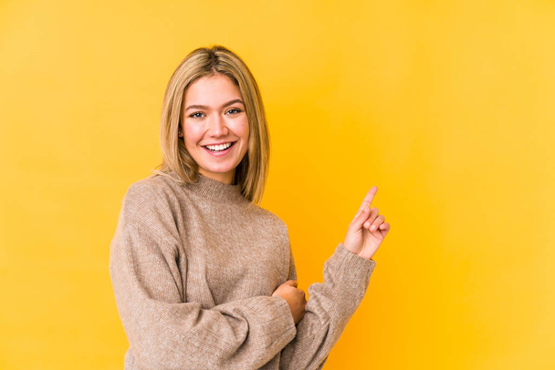 Young blonde caucasian woman isolated on yellow background smiling cheerfully pointing with forefinger away. - 写真・画像