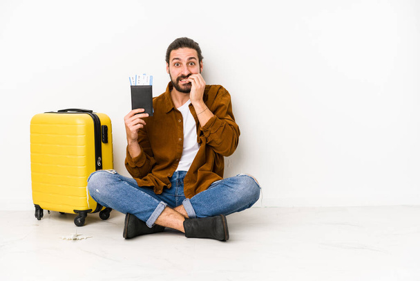 Young caucasian man sitting holding a passport and a suitcase isolated biting fingernails, nervous and very anxious. - Foto, Imagen