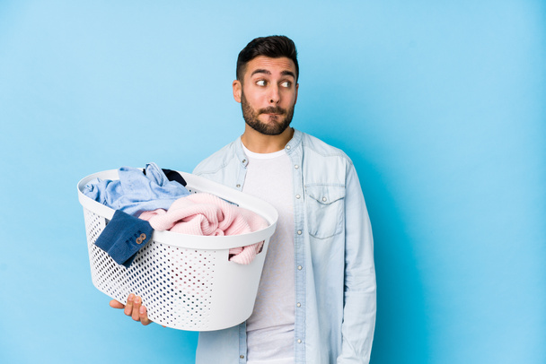 Junger gutaussehender Mann beim Wäschewaschen isoliert verwirrt, fühlt sich zweifelhaft und unsicher. - Foto, Bild