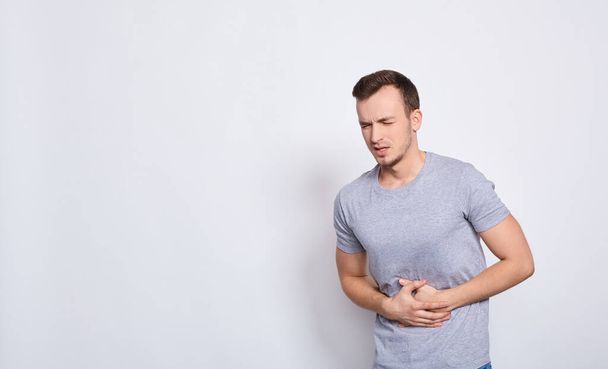 Young man in a gray t-shirt, touching his belly isolated on a white background space. Stomach ache or pain concept. Man suffering from stomach pain on a white background. Copy space. - Zdjęcie, obraz