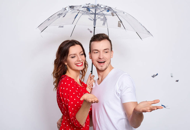 Portert, beautiful young happy couple in love together, laughing, couple hiding under an umbrella from falling confetti, hugging, catching confetti with their hands isolated on a white background. - Fotó, kép