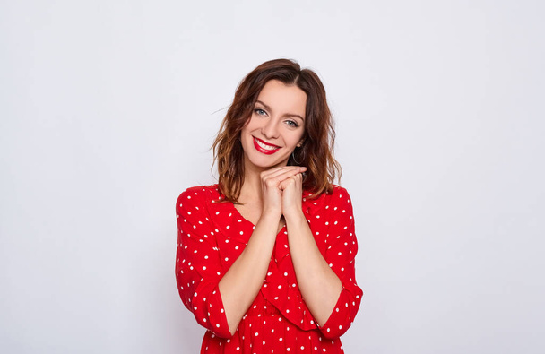 Charming cute young brown-haired woman with blue eyes and bright makeup, in a red dress in a white dot, folded her hands under the chin, smiles and looks straight posing on a white background. - Фото, изображение