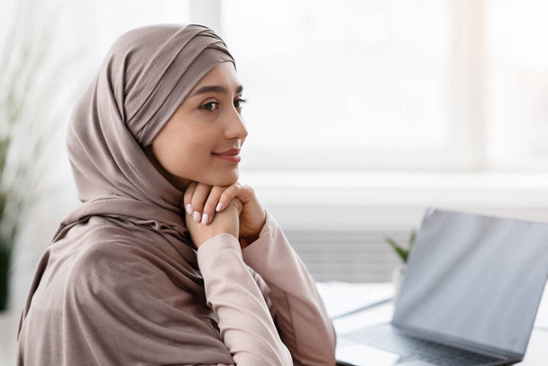 Усміхнена мусульманська бізнес-леді в хустці, сидить на робочому місці і дивиться в сторону
 - Фото, зображення
