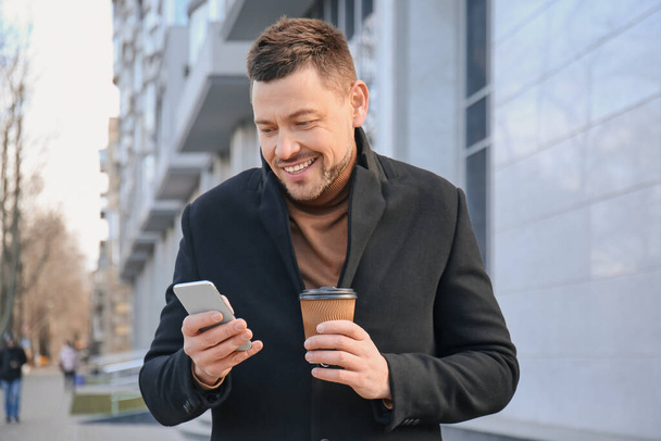 Красивый мужчина с чашкой кофе и мобильным телефоном на открытом воздухе
 - Фото, изображение
