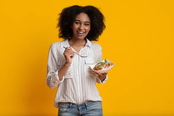 Hymyilevä afro nainen syö kasvissalaattia, seisoo keltaisen taustan päällä
 - Valokuva, kuva
