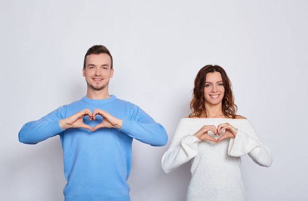 Funny happy young Caucasian lovers in light fashionable clothes, stand next to each other, show their hands heart, are in love, test emotions, smile on a white background. - Fotografie, Obrázek