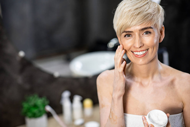 Woman Applying Cream On Face Smiling To Camera In Bathroom - Foto, Bild